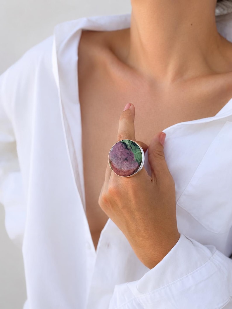 Puede incluir: Un anillo grande y redondo con una banda plateada. El anillo presenta una piedra natural con una mezcla de colores rosa, verde y blanco. El anillo se lleva en un dedo, y la persona lleva una camisa blanca abotonada.