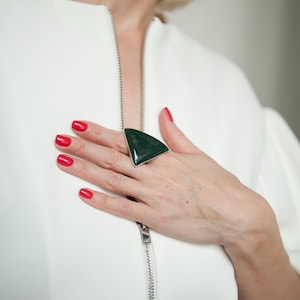 May include: A large triangular ring featuring a dark green stone set in a silver-toned metal frame. The ring is worn on a finger, positioned against a white garment with a prominent silver-colored vertical zipper running down the center.