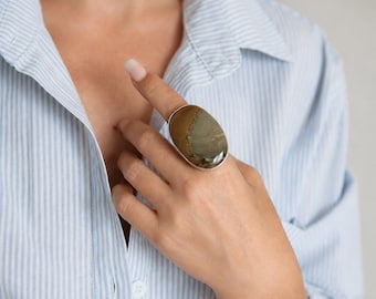 Può includere: Un grande anello statement con una pietra di diaspro ovale e lucida nei toni del marrone e del beige, incastonata in una fascia di metallo liscio color argento. La pietra presenta variazioni di colore naturali e una finitura lucida.