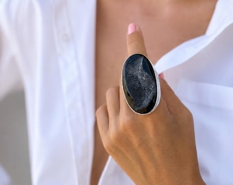 Può includere: Un anello ovale con una pietra scura e maculata incastonata in una fascia d'argento. L'anello è indossato su un dito, sullo sfondo di una camicia bianca.