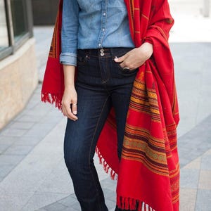 May include: A person wearing a denim shirt, dark blue jeans, and a vibrant red shawl with patterned details and fringe. The outfit is completed with pink shoes, creating a stylish and colorful ensemble.