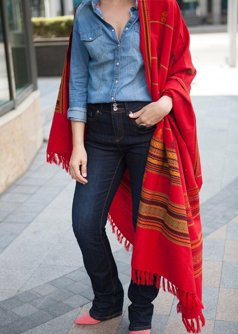 May include: A person wearing a denim shirt, dark blue jeans, and a vibrant red shawl with patterned details and fringe. The outfit is completed with pink shoes, creating a stylish and colorful ensemble.