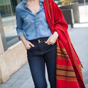 May include: A woman wearing a denim shirt, dark blue jeans, and a vibrant red shawl with a patterned border. The shawl is draped over the shoulders, adding a pop of color to the outfit. The outfit is casual and stylish.