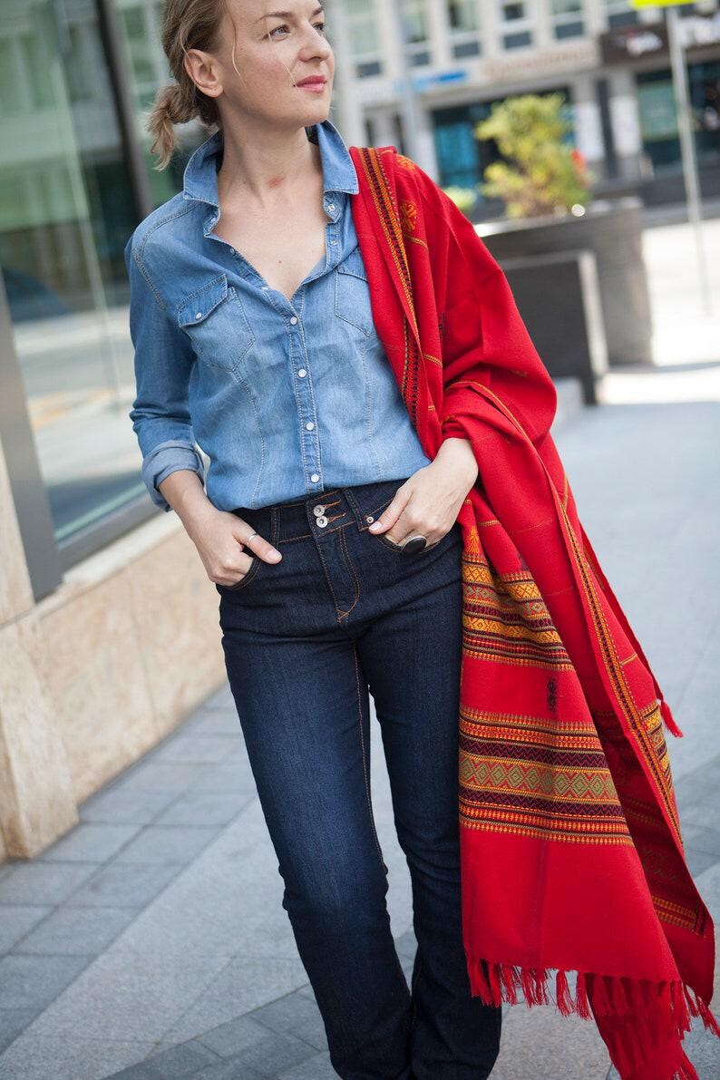 May include: A woman wearing a denim shirt, dark blue jeans, and a vibrant red shawl with a patterned border. The shawl is draped over the shoulders, adding a pop of color to the outfit. The outfit is casual and stylish.