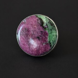 Puede incluir: Un anillo grande y redondo con una banda plateada. La piedra es una mezcla de tonos morados y verdes profundos, creando un contraste llamativo. El anillo se presenta sobre un fondo oscuro, resaltando los colores y detalles.