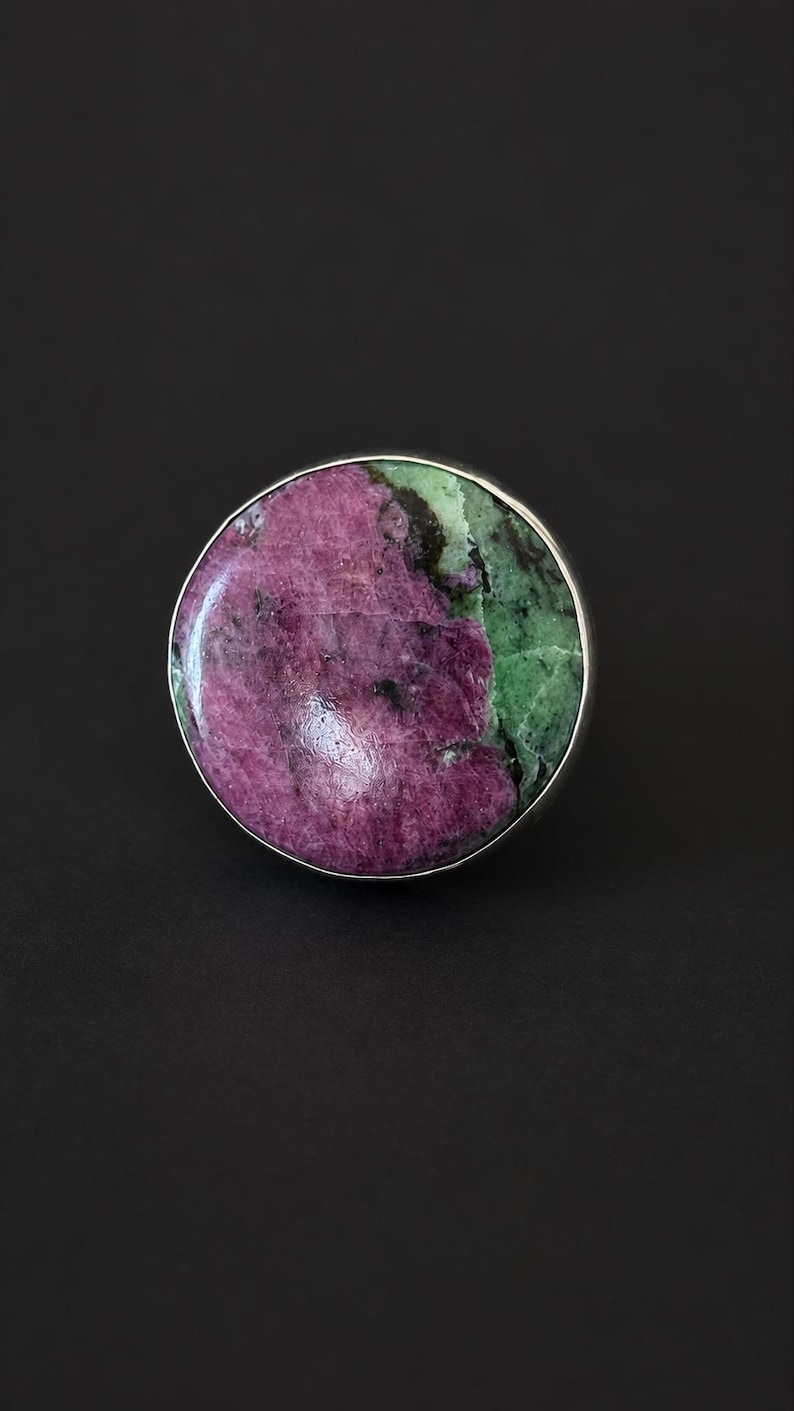 Puede incluir: Un anillo grande y redondo con una banda plateada. La piedra es una mezcla de tonos morados y verdes profundos, creando un contraste llamativo. El anillo se presenta sobre un fondo oscuro, resaltando los colores y detalles.