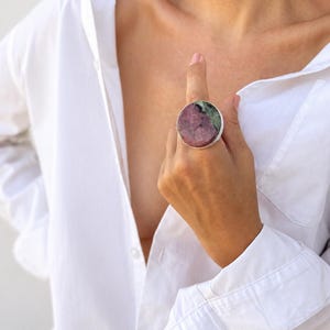 Puede incluir: Un anillo grande y redondo con una banda plateada y una piedra colorida. La piedra presenta una mezcla de tonos rosas y verdes. El anillo se lleva en un dedo y el fondo es una camisa blanca.