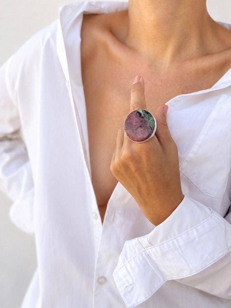 Puede incluir: Un anillo grande y redondo con una banda plateada y una piedra colorida. La piedra presenta una mezcla de tonos rosas y verdes. El anillo se lleva en un dedo y el fondo es una camisa blanca.