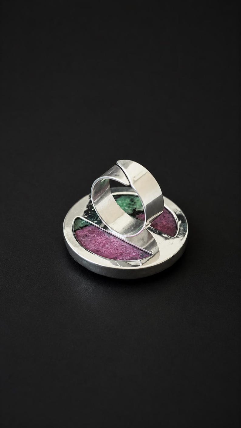 Puede incluir: Un anillo de plata con una cara grande y redonda. La cara est&aacute; incrustada con secciones de piedras verdes y magenta. La banda del anillo es una banda simple y ancha. El anillo est&aacute; sobre un fondo negro.