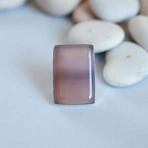 Pu&ograve; includere: Un anello rettangolare con una pietra liscia e traslucida color rosa polvere, incastonata in una sottile cornice metallica. La pietra presenta una tonalit&agrave; rosa violaceo tenue e opaca con una superficie lucida che riflette la luce.