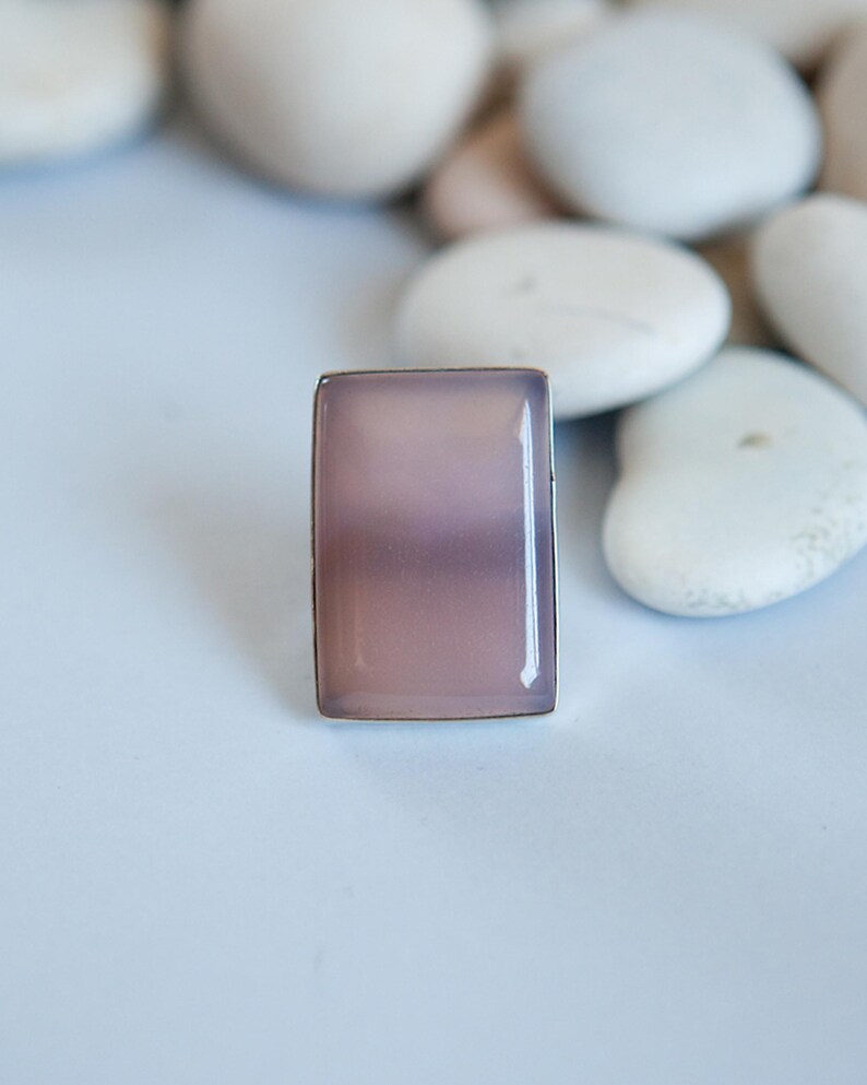 Pu&ograve; includere: Un anello rettangolare con una pietra liscia e traslucida color rosa polvere, incastonata in una sottile cornice metallica. La pietra presenta una tonalit&agrave; rosa violaceo tenue e opaca con una superficie lucida che riflette la luce.