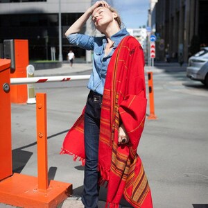 May include: A woman wearing a denim shirt and dark blue jeans, draped with a vibrant red shawl featuring intricate striped patterns in gold and brown. The shawl has fringed edges. Pink shoes complete the outfit.