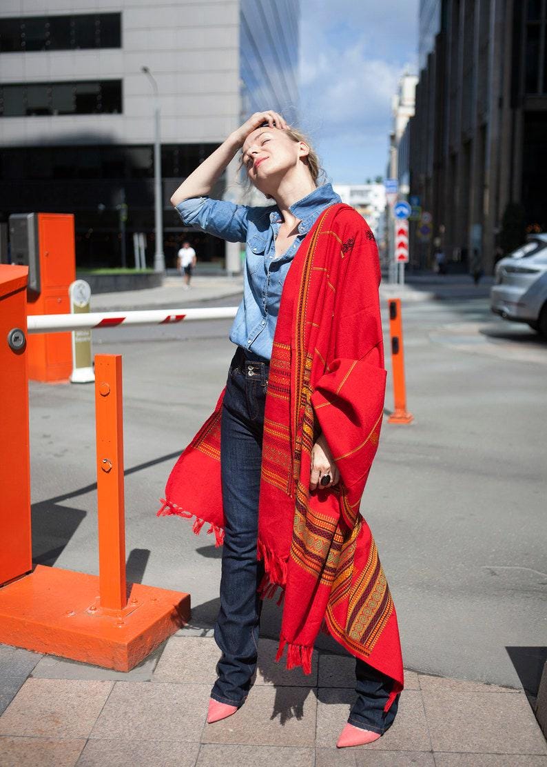 May include: A woman wearing a denim shirt and dark blue jeans, draped with a vibrant red shawl featuring intricate striped patterns in gold and brown. The shawl has fringed edges. Pink shoes complete the outfit.