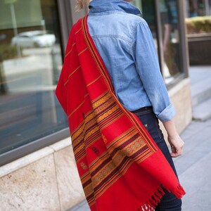 May include: A vibrant red shawl with intricate geometric patterns in gold, brown, and green. The shawl has a fringed edge and is draped over the shoulder. The textile is a bold statement piece.