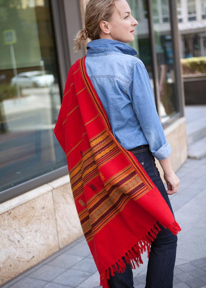 May include: A vibrant red shawl with intricate geometric patterns in gold, brown, and green. The shawl has a fringed edge and is draped over the shoulder. The textile is a bold statement piece.