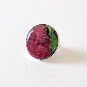 Puede incluir: Un anillo redondo con una gema grande y pulida. La piedra se divide en dos secciones: un tono rojo-rosa intenso y un verde vibrante. La banda del anillo es de plata pulida.