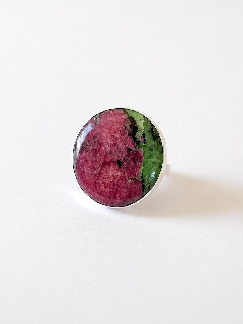 Puede incluir: Un anillo redondo con una gema grande y pulida. La piedra se divide en dos secciones: un tono rojo-rosa intenso y un verde vibrante. La banda del anillo es de plata pulida.