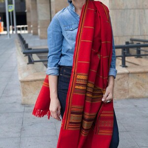 May include: A vibrant red scarf with intricate geometric patterns in gold, orange, and green. The scarf has a fringed edge and is draped over the shoulders. The scarf is a textile accessory.