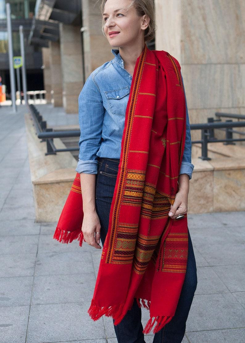 May include: A vibrant red scarf with intricate geometric patterns in gold, orange, and green. The scarf has a fringed edge and is draped over the shoulders. The scarf is a textile accessory.