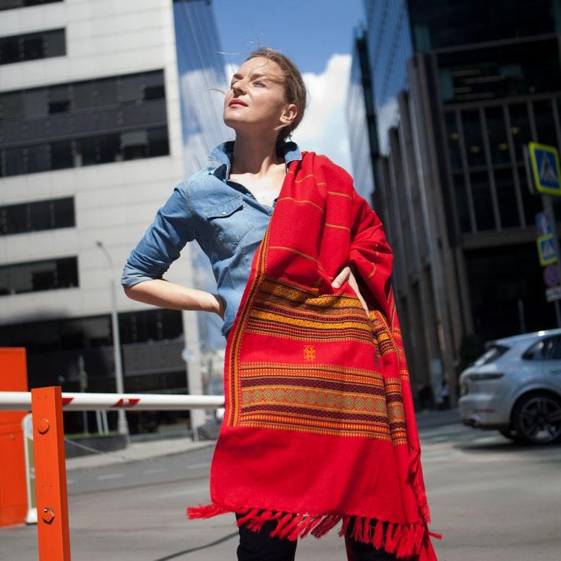 May include: A vibrant red shawl with a detailed pattern in gold and brown. The shawl has a fringed edge and is draped over the shoulders. The person is wearing a denim shirt.