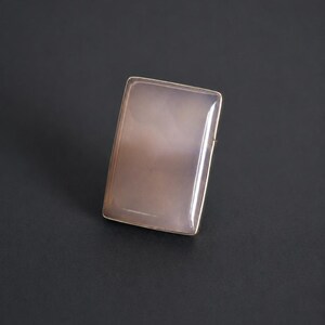 Pu&ograve; includere: Un anello rettangolare con una pietra liscia e lucida di colore grigio rosato pallido, incastonata in una sottile cornice metallica. La pietra ha una qualit&agrave; traslucida con sottili variazioni interne nuvolose e un design semplice.