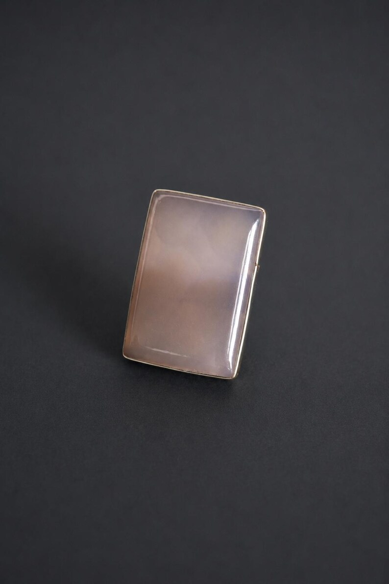 Pu&ograve; includere: Un anello rettangolare con una pietra liscia e lucida di colore grigio rosato pallido, incastonata in una sottile cornice metallica. La pietra ha una qualit&agrave; traslucida con sottili variazioni interne nuvolose e un design semplice.