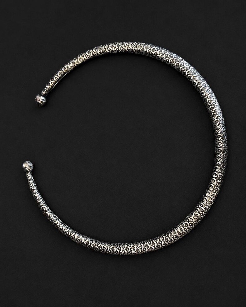Peut inclure: Un collier ras du cou argent&eacute; avec un motif textur&eacute; en forme d'&eacute;cailles. Le collier a une forme arrondie avec une petite boule &agrave; chaque extr&eacute;mit&eacute;. Le fond est noir uni.