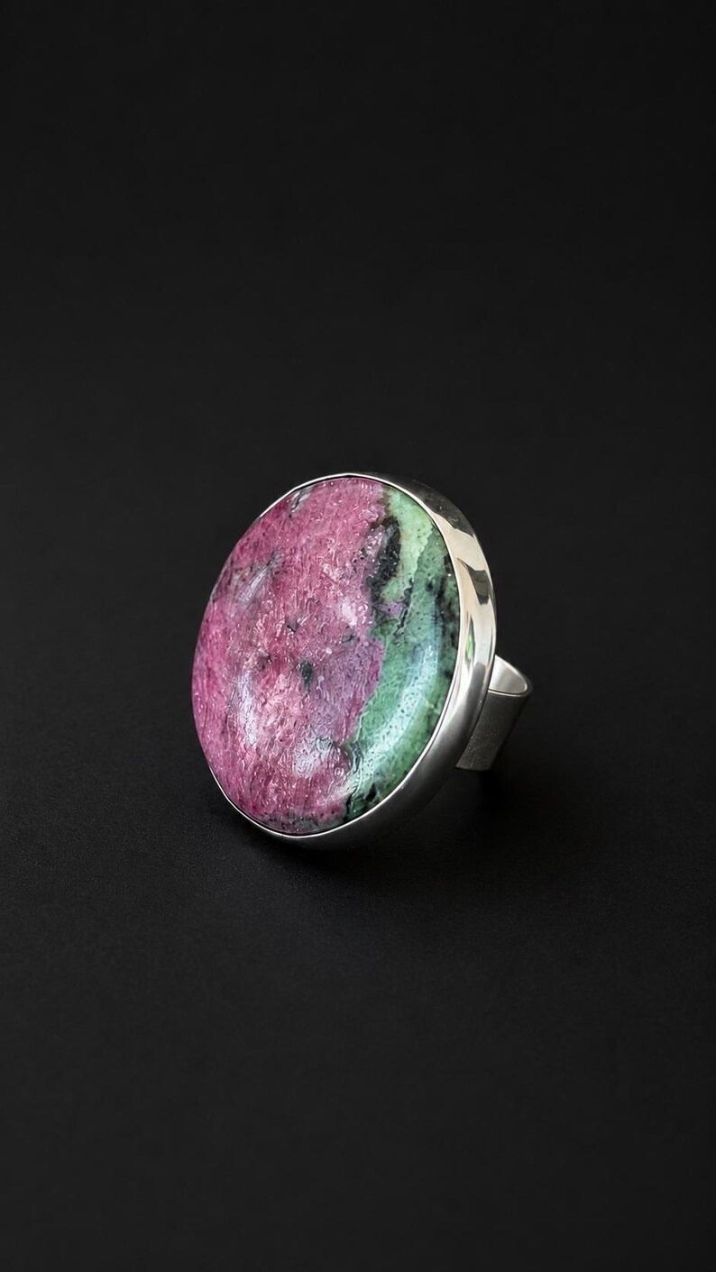 Puede incluir: Anillo llamativo con una piedra preciosa ovalada pulida en tonos rosa, verde y negro, engastada en una ancha banda de plata pulida. El anillo se muestra sobre un fondo negro. Una joya audaz.