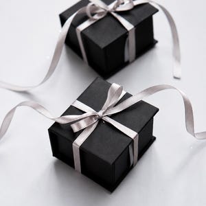Pu&ograve; includere: Due scatole regalo quadrate nere, ciascuna caratterizzata da una finitura opaca liscia e un coperchio incernierato. Un nastro di raso grigio chiaro &egrave; legato a fiocco attorno al centro di ogni scatola, con le estremit&agrave; che si estendono.
