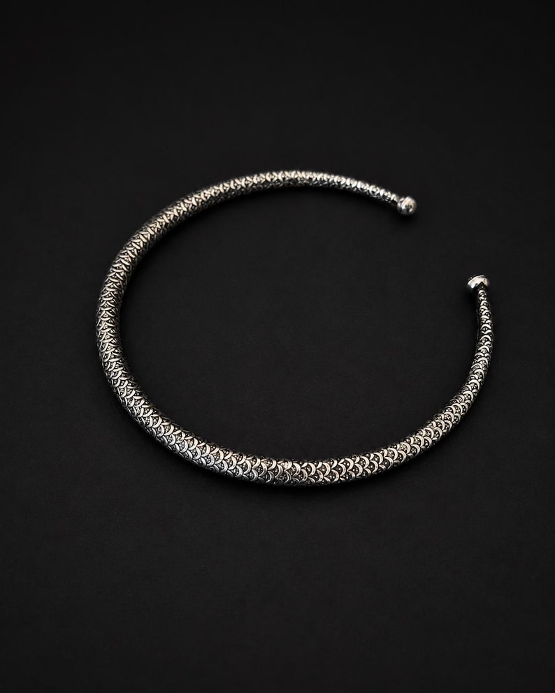 Peut inclure: Un collier ras du cou argent&eacute; avec un motif textur&eacute; et d&eacute;taill&eacute;. Le collier a une forme arrondie et se r&eacute;tr&eacute;cit aux extr&eacute;mit&eacute;s, avec de petites fermetures en forme de boule. Le fond est noir uni.