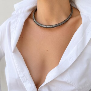 Peut inclure: Un collier argent&eacute; avec un motif r&eacute;p&eacute;titif est port&eacute; sur une chemise blanche boutonn&eacute;e. Le collier est de style ras du cou, reposant pr&egrave;s du cou. La chemise est d&eacute;boutonn&eacute;e en haut.