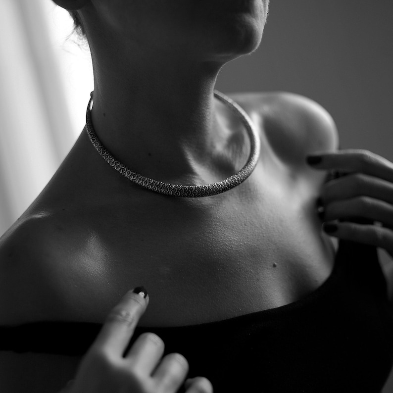 Peut inclure: Un collier ras du cou argent&eacute; avec un motif textur&eacute; et d&eacute;taill&eacute;. Le collier est port&eacute; autour du cou, et l'image est en noir et blanc. La personne porte un haut noir.