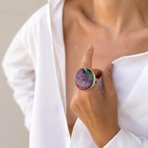 Puede incluir: Un anillo llamativo con una piedra preciosa grande y redonda en tonos rosa, verde y blanco, engastada en una banda plateada. El anillo es una pieza de joyer&iacute;a audaz, adecuada para moda o como elemento decorativo.
