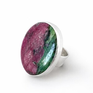 Puede incluir: Anillo de plata con una piedra preciosa grande y ovalada. La piedra preciosa muestra una mezcla de colores, incluyendo tonos rosa intenso y verde. El anillo tiene una banda de plata pulida.