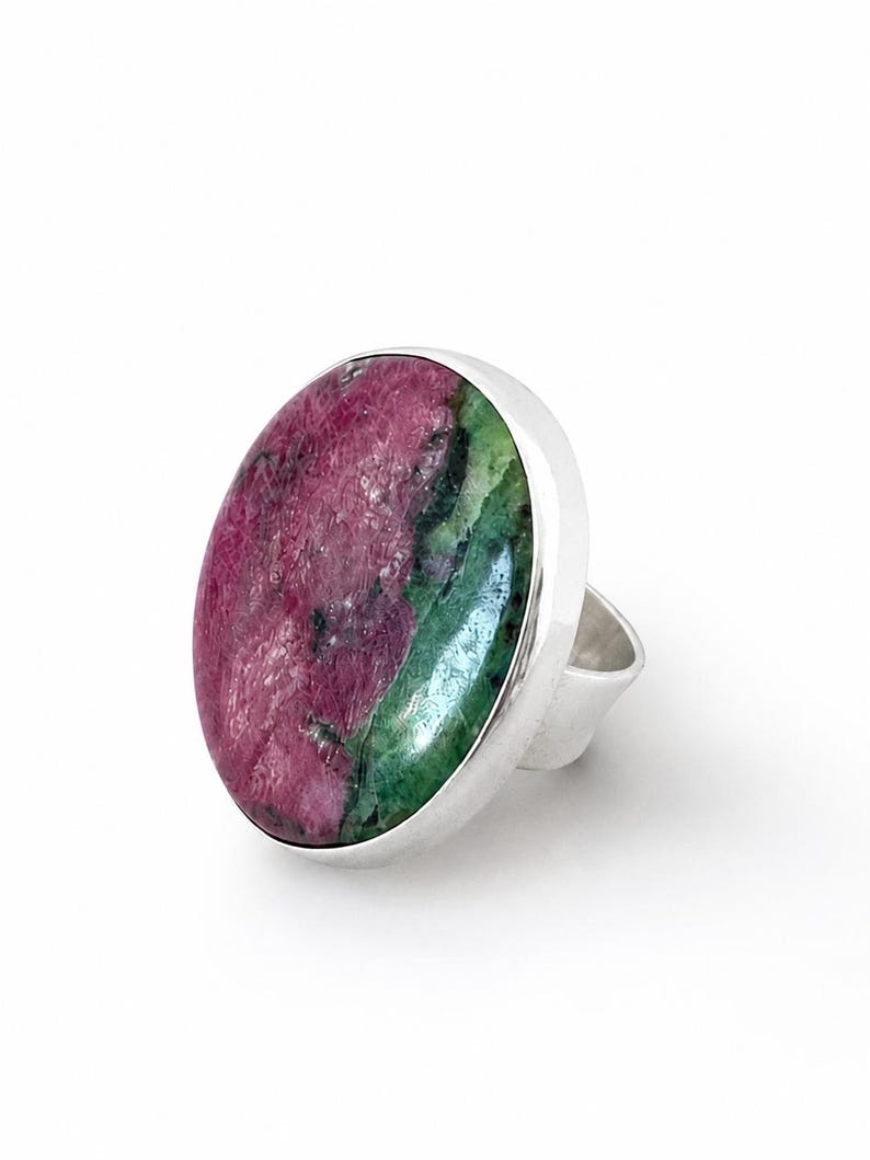 Puede incluir: Anillo de plata con una piedra preciosa grande y ovalada. La piedra preciosa muestra una mezcla de colores, incluyendo tonos rosa intenso y verde. El anillo tiene una banda de plata pulida.