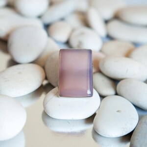 Pu&ograve; includere: Un anello rettangolare con una pietra liscia, traslucida e di colore viola chiaro incastonata in una semplice fascia metallica. La pietra presenta una superficie piatta con bordi smussati che riflettono la luce sulla tonalit&agrave; lavanda.