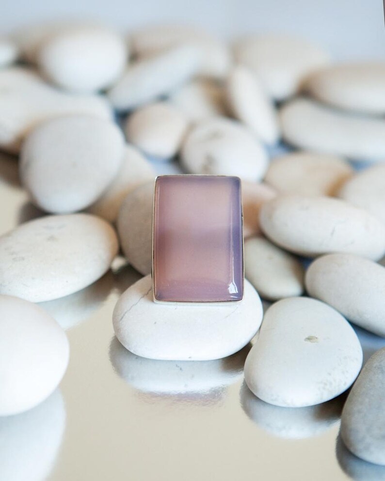 Pu&ograve; includere: Un anello rettangolare con una pietra liscia, traslucida e di colore viola chiaro incastonata in una semplice fascia metallica. La pietra presenta una superficie piatta con bordi smussati che riflettono la luce sulla tonalit&agrave; lavanda.