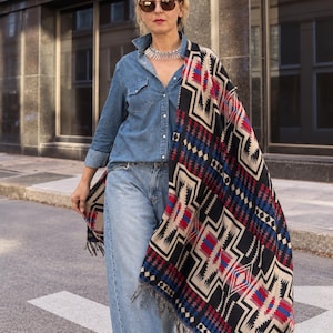 May include: A blue denim button-down shirt, light blue denim jeans, and a large black, red, and cream patterned shawl with fringe edges. The shawl features a geometric design. A silver-toned metal collar necklace and brown sunglasses are worn.