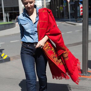 May include: A woman wearing a denim shirt and dark blue jeans, holding a vibrant red shawl with intricate patterns and fringe. The shawl features a geometric design in shades of yellow, orange, and brown. The scene is set on a city street.
