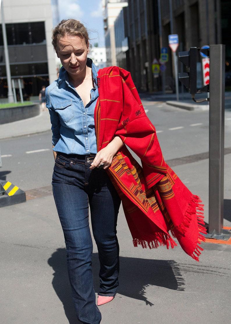 May include: A woman wearing a denim shirt and dark blue jeans, holding a vibrant red shawl with intricate patterns and fringe. The shawl features a geometric design in shades of yellow, orange, and brown. The scene is set on a city street.