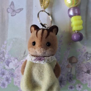 May include: A brown and tan chipmunk figurine wearing a light yellow dress with a white lace collar. The chipmunk is attached to a keychain with a beaded strand of yellow, purple, and iridescent beads.