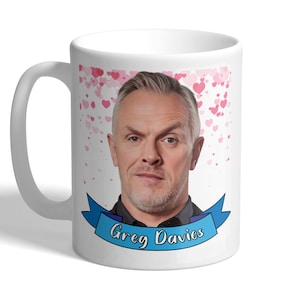 May include: White ceramic mug featuring a portrait of Greg Davies with a blue banner that reads "Greg Davies". The mug has a background of pink hearts.