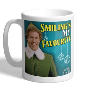 May include: White ceramic mug with a smiling man in a green elf costume. The mug has the text "Smiling's My Favourite" and the quote "-Buddy The Elf" on a teal background with hanging baubles.