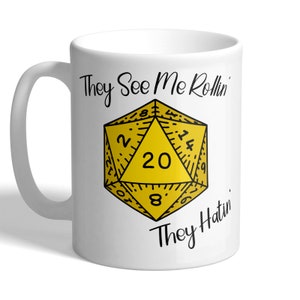 May include: White ceramic mug featuring a black and yellow illustration of a twenty-sided die. The text "They See Me Rollin' They Hatin'" is printed on the mug.
