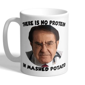 May include: White ceramic mug with a portrait of a man wearing glasses and a stethoscope. The mug has the text "THERE IS NO PROTEIN IN MASHED POTATO" printed in black.