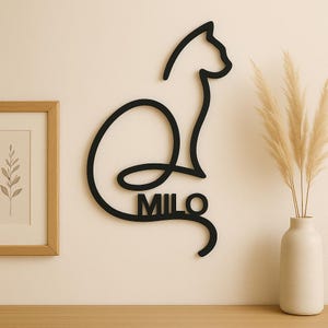 Peut inclure: Décoration murale noire en forme de chat avec le nom "MILO". Le chat est représenté par un trait continu, créant une silhouette minimaliste. L'œuvre est fixée sur un mur neutre, avec un cadre et un vase de pampas en arrière-plan.