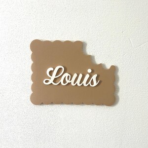 Peut inclure: Une plaque en forme de biscuit, de couleur marron et festonnée, avec le nom "Louis" écrit en lettres blanches en cursive.