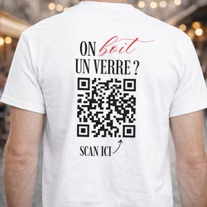 Peut inclure: T-shirt blanc avec texte noir "ON boit UN VERRE?" dans une police stylisée, avec un code QR en dessous et les mots "SCAN ICI" avec une flèche pointant vers le code QR. Le t-shirt est uni.
