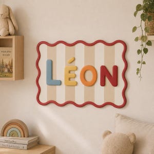Op de afbeelding: Een rechthoekig wandbord met een golvende rode rand en een beige en wit gestreepte achtergrond. De naam LÉON wordt weergegeven in verhoogde, driedimensionale letters in blauw, geel, oranje en rood, met een accentteken op de letter E.