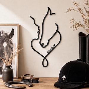 Peut inclure: Une décoration murale en métal noir représentant un dessin au trait continu d'un profil humain et d'une tête de cheval formant un cœur, avec le texte "EQUID.IY" intégré au design. L'objet est fixé sur un mur de couleur claire.